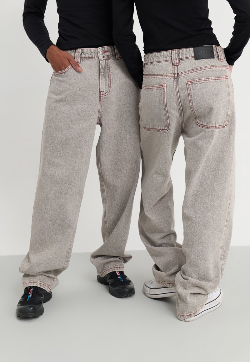 Vaqueros gris claro con un corte holgado, costuras rojas visibles y dos bolsillos traseros. Usados con zapatillas en blanco y negro.