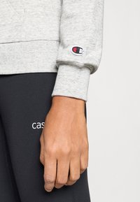 Κοντινό ενός χεριού που ξεκουράζεται στο πλάι, φορεμένο με ανοιχτό γκρι φούτερ Champion και μαύρο παντελόνι με μερικό λευκό κείμενο "cas".