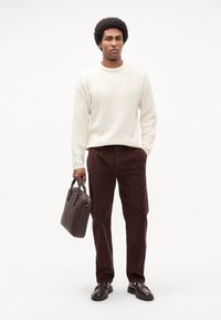 Lys beige ribbet sweater, brune fløjlsbukser, sorte loafers, holder en mørkebrun lædertaske, sat mod en ensfarvet hvid baggrund.