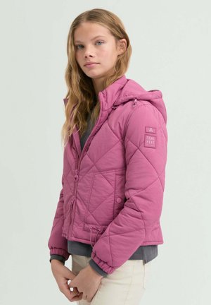 Mujer joven con chaqueta acolchada rosa con capucha, de pie con las manos ligeramente entrelazadas, contra un fondo claro y liso.