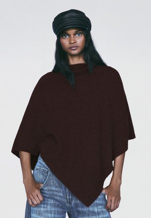 Femme portant une casquette en cuir noire, un pull poncho marron foncé et un jean bleu, debout les mains dans les poches devant un fond uni.