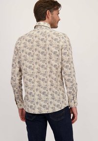 Man in een beige bloemenoverhemd met lange mouwen en donkerblauwe jeans, staande met zijn rug naar de camera tegen een effen lichte achtergrond.