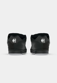 Schwarze Sneaker mit glatter Textur, runder Zehenpartie und kontrastierendem weißen Logo-Detail an der Ferse. Gummisohle mit sichtbaren Nahtakzenten.