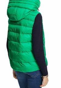 Veste sans manches matelassée verte avec capuche portée par-dessus un pull texturé marine à manches longues et un jean en denim bleu, vue de dos.