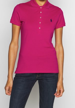 Person iført en tætsiddende magentarød kortærmet poloshirt med knapper og mørkeblå jeans, armene afslappet langs siderne.