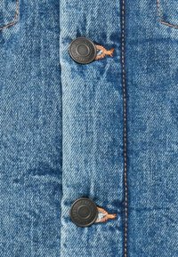 Denimskjorta med en ljusblå textur, två svarta knappar, orange sömnad och vertikal sömdetaljering. Närbild av tyget.