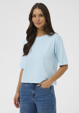 Kobieta z długimi brązowymi włosami ubrana w jasnoniebieską bluzkę z krótkim rękawem i niebieskie dżinsy, z jedną ręką w kieszeni na jednolitym tle.