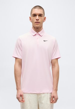 Junger Mann mit einem hellrosa Nike-Poloshirt und hellen gemusterten Shorts, stehend vor einem weißen Hintergrund.