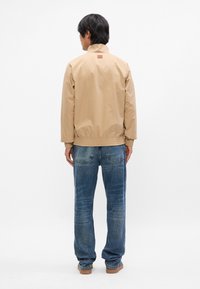 Beige ελαφρύ μπουφάν με ψηλό γιακά, ελαστικούς μανσέτες και τελείωμα. Φορεμένο πάνω από μπλε τζιν παντελόνι, αναδεικνύει μια άνετη εφαρμογή.