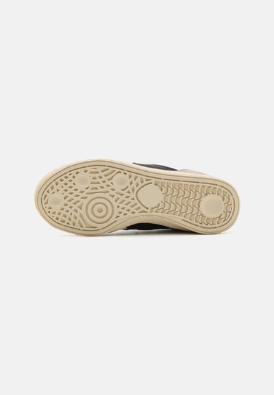 Black shoe with a textured beige rubber sole, featuring a patterned tread for grip. The sole has a rounded shape with various embossed details.