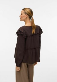 Blouse marron à volants avec une coupe décontractée, dotée de manches longues et d'un design en couches, associée à un pantalon beige à jambes larges.