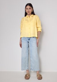 Camicia gialla in lino con bottoni e due tasche frontali, abbinata a jeans in denim a gamba larga di colore blu chiaro e sandali scivolati color tan.