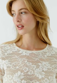 Lace blouse in off-white with floral patterns, round neckline, and sheer texture, featuring a delicate dot design throughout.