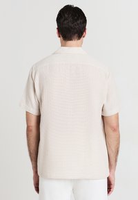 Camicia corta manica color beige chiaro con un tessuto texturizzato e un delicato motivo intrecciato, caratterizzata da un colletto rilassato e linee semplici e pulite.
