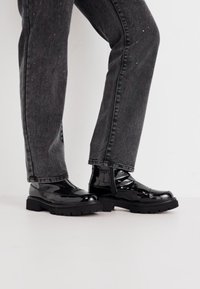 Bottines en patent noir avec une semelle épaisse, comportant des accents de sangle. Portées avec un jean gris foncé, affichant une coupe slim et de légères embellissements.