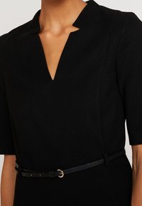 Schwarzes Kleid mit V-Ausschnitt, kurzen Ärmeln, strukturiertem Stoff und einem schmalen schwarzen Gürtel mit einer goldenen Ring-Schnalle.