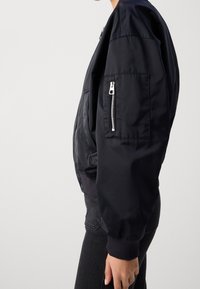 Černá bomber bunda s hladkým, lesklým povrchem, žebrovanými manžetami a zipovou kapsou na levém rukávu. Navržena s uvolněnou siluetou.