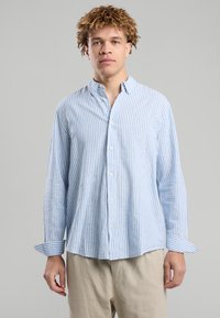 Young man with curly hair wearing a light blue and white vertical striped button-up shirt and beige pants against a neutral background.