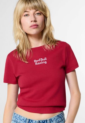 Jonge vrouw met blond haar draagt een rood cropped t-shirt met de tekst "Red Bull Racing" en lichtblauwe spijkerbroek tegen een effen achtergrond.