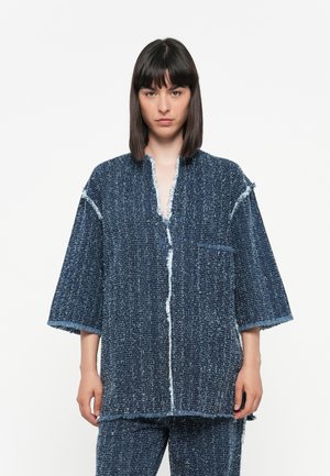 Veste texturée bleue avec une coupe décontractée, présentant un col en V, des manches courtes, des bords effilochés et une poche poitrine unique.