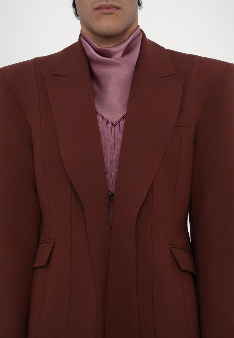 Brauner maßgeschneiderter Blazer mit scharfen Revers, Fronttaschen und strukturierten Schultern. Darunter ein rosa Satinschal und ein fransiger, strukturierter Strickoberteil.