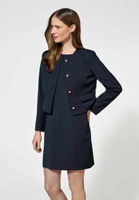 Granatowa marynarka o dopasowanym kroju z złotymi guzikami, zestawiona z pasującą sukienką. Gładki, strukturalny fason, długie rękawy i gładka tekstura.