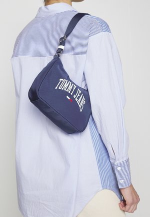 Person wearing blue and white striped shirt carrying navy blue Tommy Jeans shoulder bag across back.