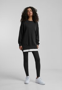 Black sweatshirt dress with long sleeves, side slits, and a white layered hem, paired with black leggings and white sneakers. Smooth texture.