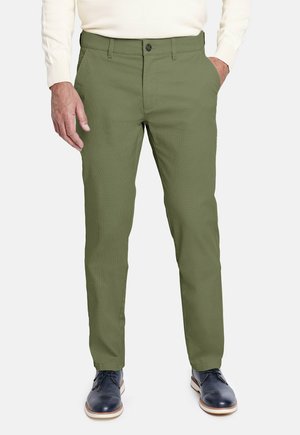 Hombre que lleva pantalones ajustados verde oliva, una camisa de manga larga blanca y zapatos de cuero azul oscuro, de pie con las manos en los bolsillos.