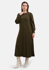 Langärmliges, olivgrünes Kleid mit vertikalen Nadelstreifen. Weicher Stoff, A-Linien-Silhouette, knielang, kombiniert mit weißen Stiefeletten mit Absatz.