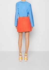 Chemise bleue texturée raccourcie, jupe en denim orange vif, chaussures à plateforme jaunes et à fleurs, présentées de dos sur un fond uni.