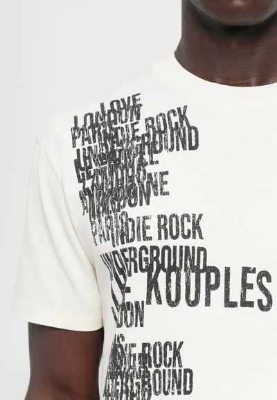 Λευκό βαμβακερό μπλουζάκι με μαύρη, φθαρμένη εκτύπωση κειμένου που περιλαμβάνει διάφορες λέξεις όπως "ΑΓΑΠΗ", "ΡΟΚ" και "ΚΟΥΠΕΛΣ" τοποθετημένες σε διαγώνια διάταξη.