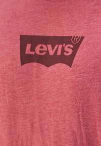 T-shirt en coton rouge avec un logo Levi's bordeaux foncé dans un design graphique stylisé sur un fond texturé.