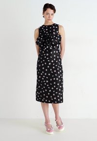 Zwarte satijnen jurk met een strikdetail; heeft een bloempatroon in roze en wit, knie-lang, aansluitende silhouet, en mouwloos ontwerp.