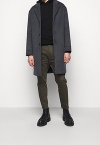 Cappotto grigio in lana sopra un hoodie nero, abbinato a pantaloni verde oliva e stivali neri con lacci. Presenta una chiusura a bottoni e una vestibilità aderente.