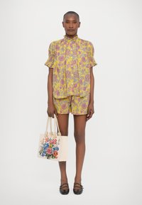 Gul blomstret skjorte og shortssett med puffede ermer, paret med en kremfarget totebag med fargerik blomsterbroderi. Svarte flate sko.