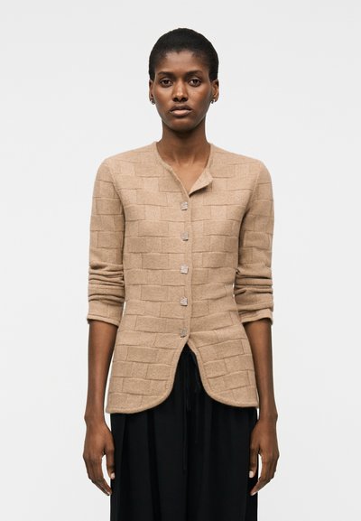 Femme portant un cardigan beige texturé boutonné avec les manches retroussées et un pantalon noir, debout devant un fond blanc uni.