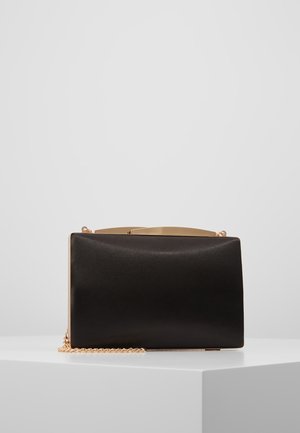 Pochette rectangulaire noire avec cadre doré et bandoulière chaîne, posée sur un piédestal blanc sur fond gris.