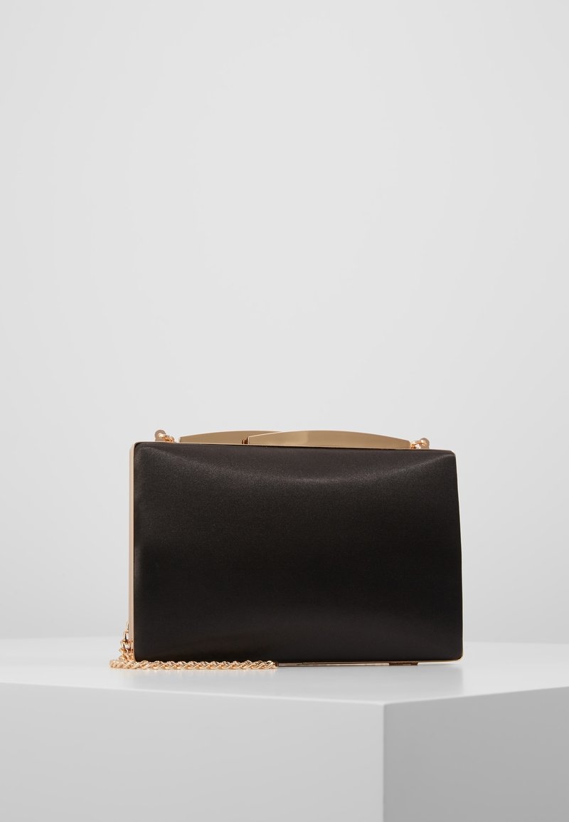 Pochette rectangulaire noire avec cadre doré et bandoulière chaîne, posée sur un piédestal blanc sur fond gris.