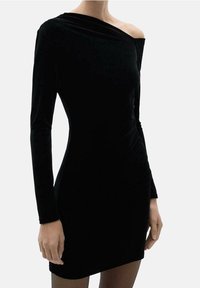 Robe en velours noir à manches longues, décolleté asymétrique et silhouette ajustée. Texture douce avec des détails froncés sur un côté.
