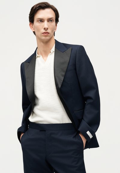 Tânăr îmbrăcat într-un costum bleumarin cu rever de satin negru și o cămașă albă texturată cu guler în V, mâinile în buzunare, privind într-o parte.