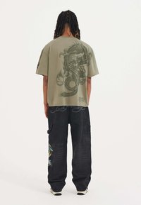Olive green oversized T-shirt featuring a large gray dragon graphic on the back. Paired with black pants displaying colorful embroidery.