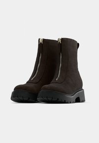 Brown suede ankle boots with a thick black rubber sole and dual silver side zippers. Features a rounded toe and a pull tab at the back.