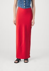 Jupe maxi rouge à pois fabriquée en tissu lisse, associée à un gilet en denim court et des sandales à talons ouverts noires.