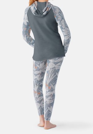Femme portant un sweat à capuche gris et des leggings assortis à motifs floraux, debout pieds nus, de dos, sur un fond uni.