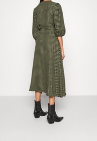 Robe midi à carreaux verts et noirs avec des manches bouffantes et une taille cintrée, associée à des bottines noires. Tissu lisse et silhouette fluide.