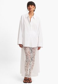 Hvid oversized skjorte med lange ærmer, parret med gennemsigtige hvide blondebukser med blomster mønstre og sorte hælede sandaler.