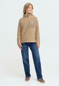 Pull en polaire beige avec un col zippé à mi-hauteur, une poche poitrine avec rabat et deux poches latérales ; associé à un jean bleu et des chaussures brunes.