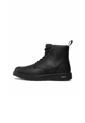 Botín negro de piel hasta el tobillo con diseño de cordones, cuello acolchado, detalles texturizados y suela Vibram con un logo amarillo.