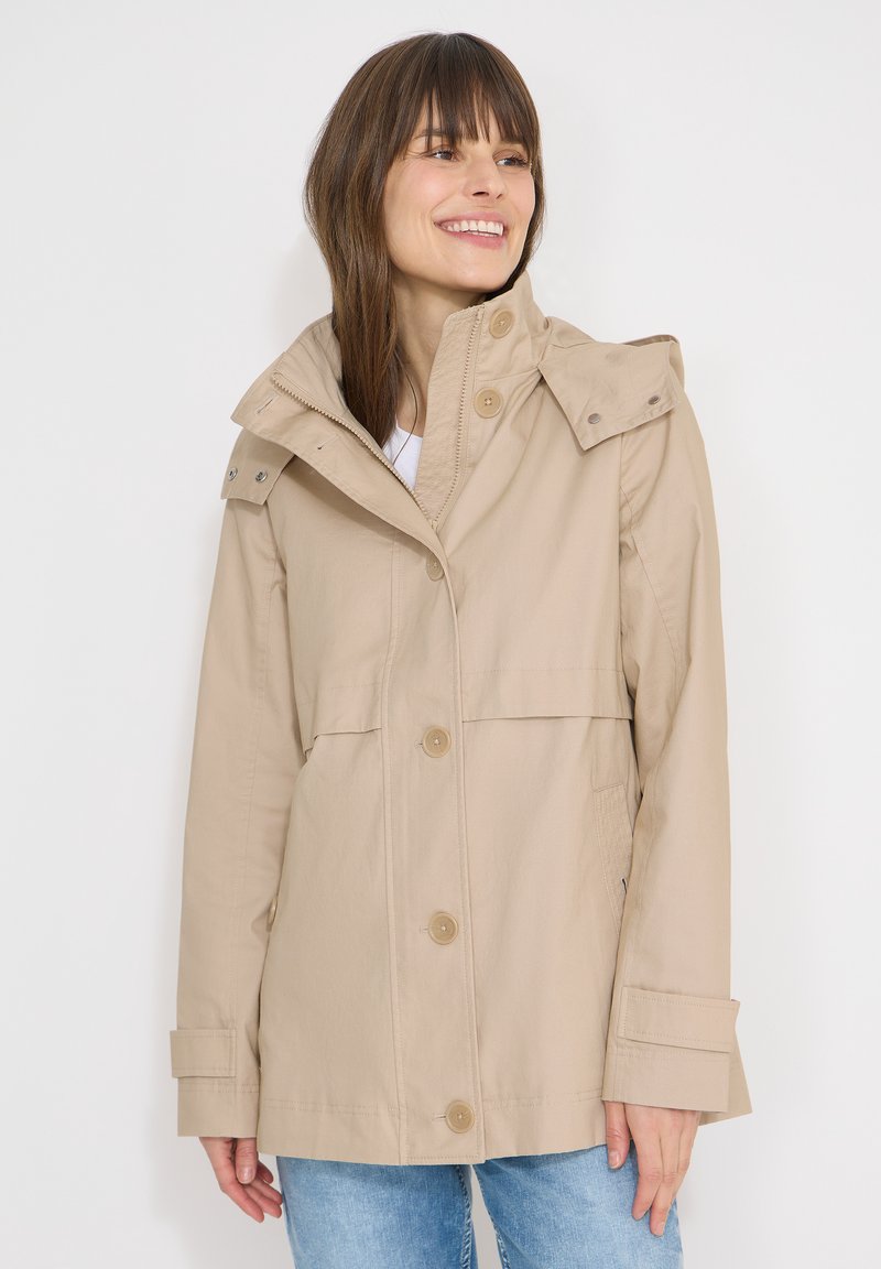 Femme souriante portant une veste à capuche beige avec fermeture par boutons et fermeture éclair, avec un jean bleu clair, sur un fond uni.